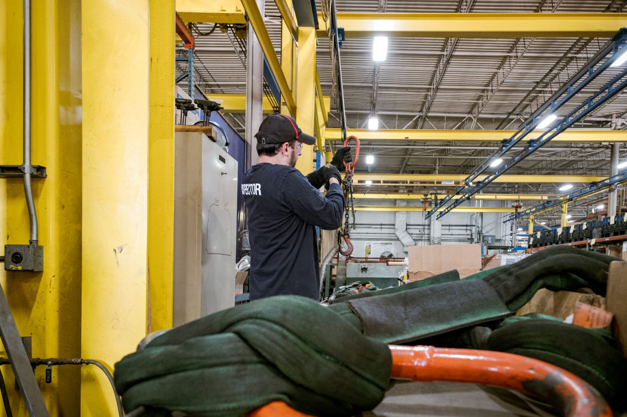 An inspector holding an alloy steel 2-leg bridle chain sling in a factory setting with used synthetic slings in the foreground.
